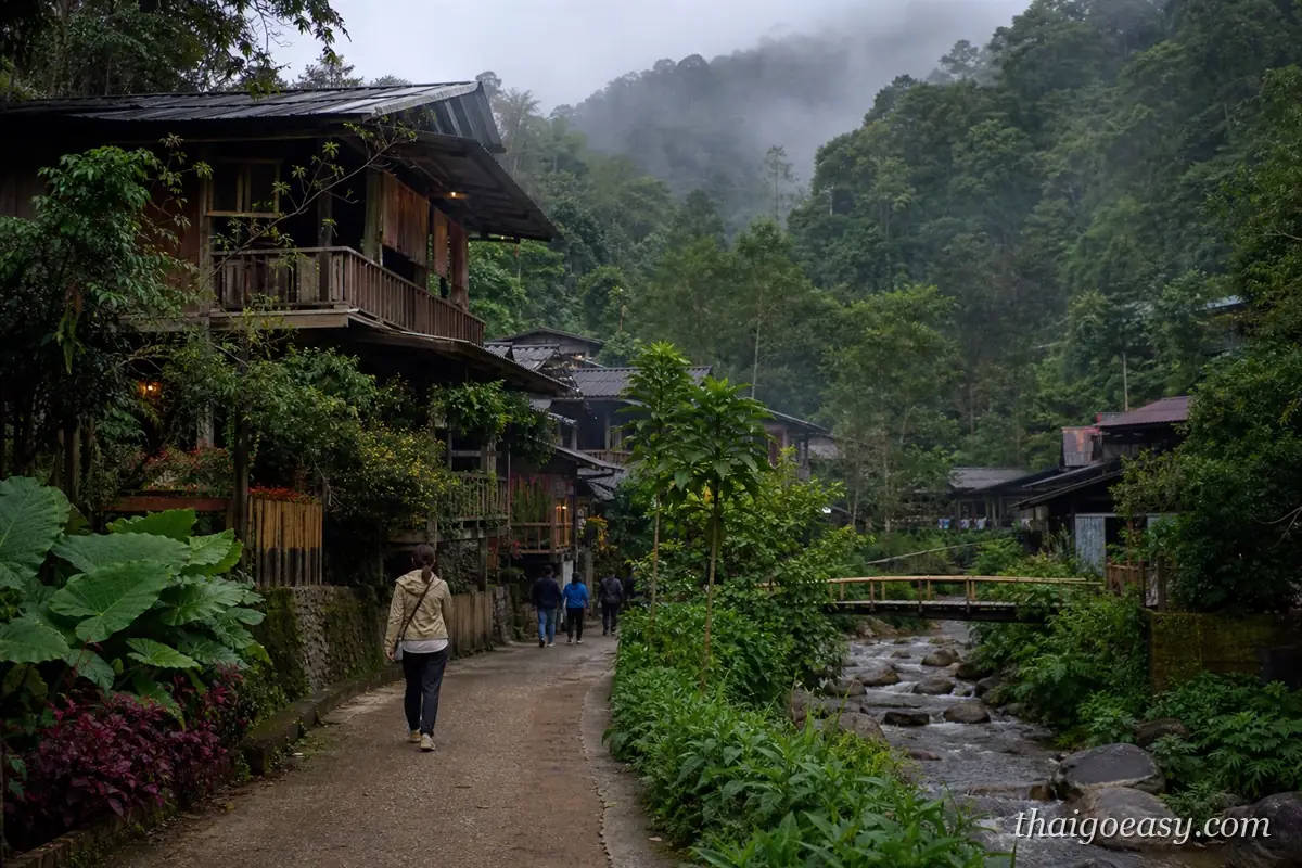 湄康蓬村的溪邊步道，旅客沿石徑往山裡走，兩側是高腳木屋與濃密森林，遠處山嵐未散
