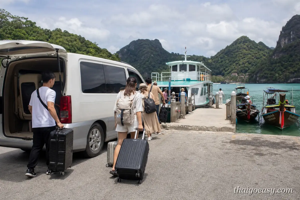 旅客拖著行李從白色接駁廂廂型車走向碼頭，前方停著渡輪與長尾船