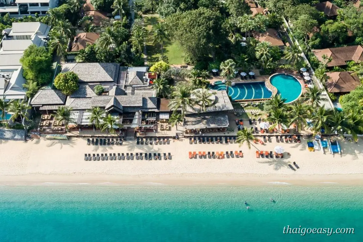 空拍 Bandara Spa Resort & Pool Villas Samui，紅褐色泰式屋頂建築沿著海灘排列，沙灘上兩排躺椅，後方是弧形泳池