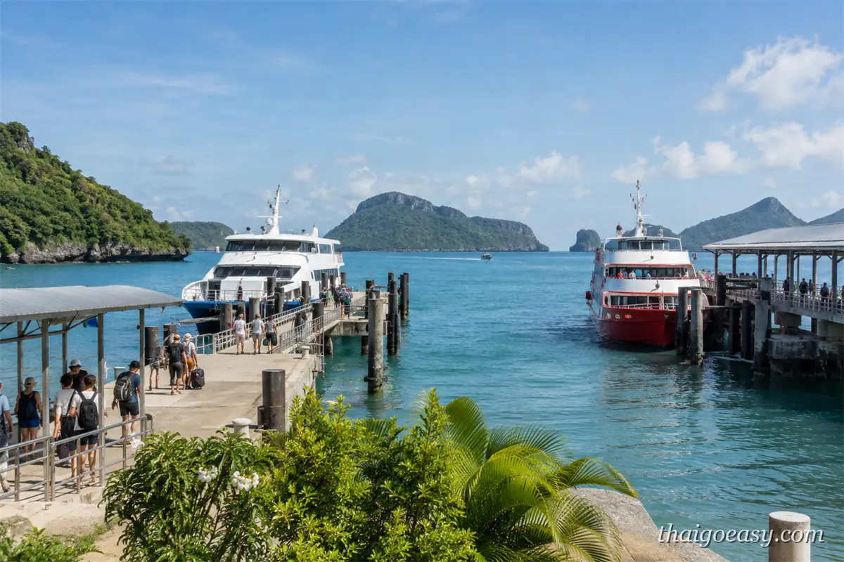 泰國島嶼碼頭同時停靠白色與紅色兩艘渡輪，旅客拖著行李走在棧橋上，遠方是多座小島