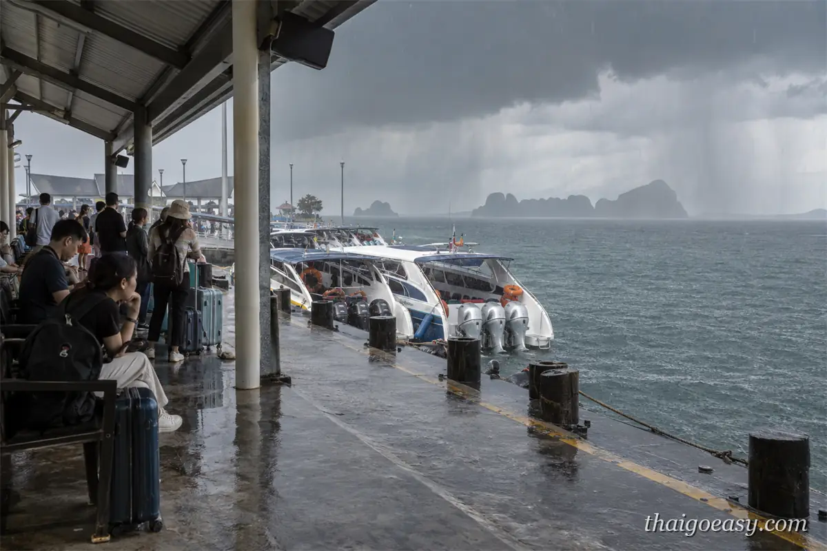 旅客在碼頭的遮雨棚下坐著等船，地面濕滑，旁邊停著數艘快艇，海面遠方是烏雲與落下的雨幕
