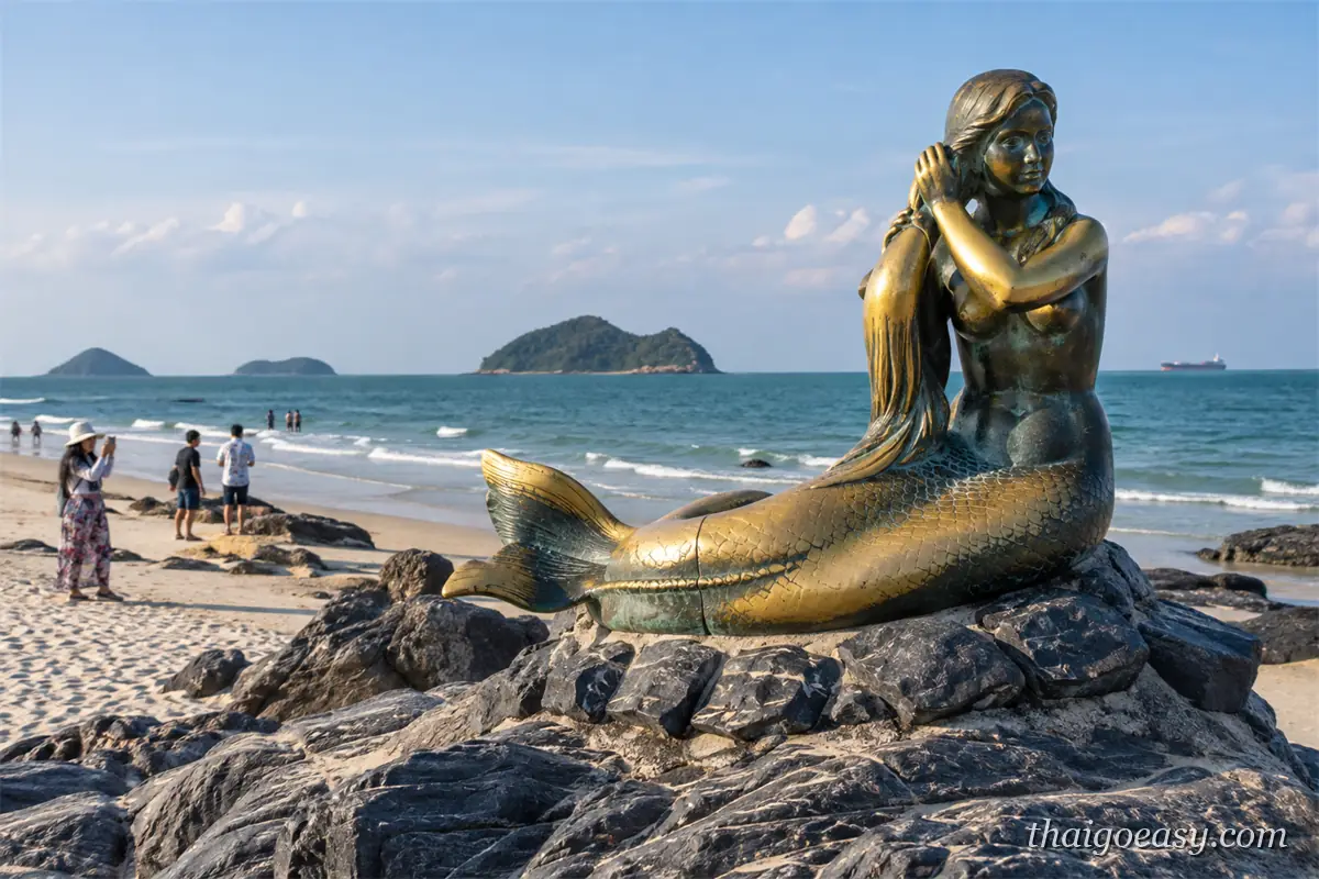 金色的美人魚銅像坐在海邊岩石上梳理長髮，背後是沙灘、遊客與海面上的小島