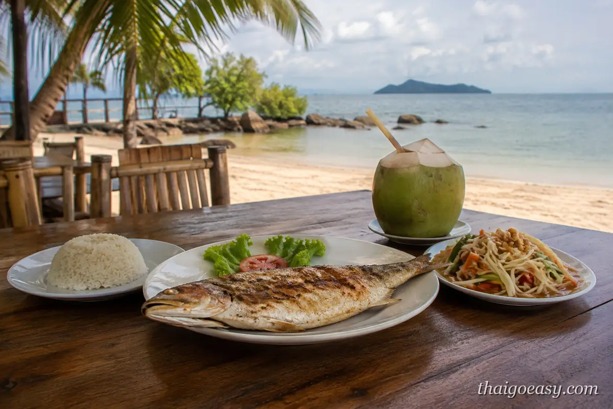 海邊木桌上的泰式餐點：烤魚、青木瓜沙拉、白飯與椰子水，背景是沙灘與遠處小島