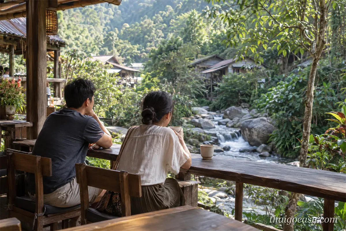 兩名旅客坐在湄康蓬溪邊咖啡店的吧台座喝咖啡，望著石灘溪流與對岸的木造村屋