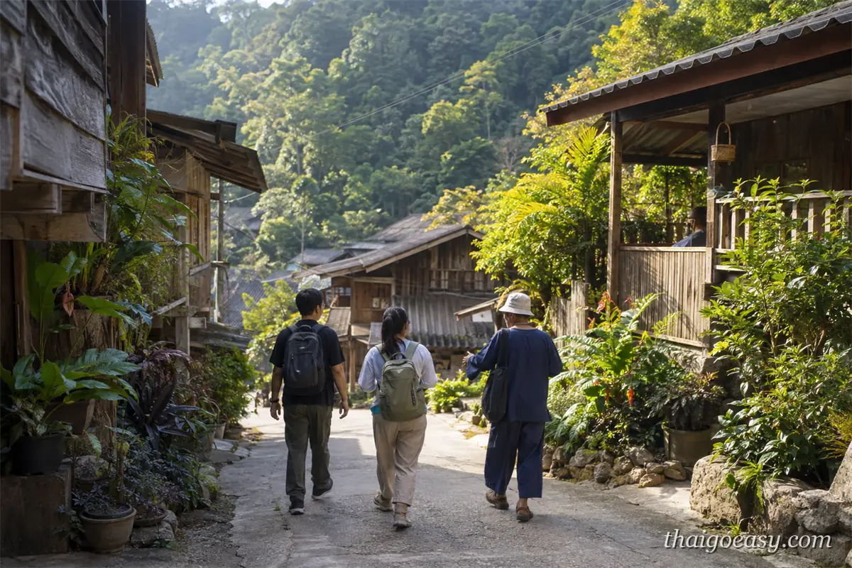 三名旅客沿著湄康蓬村的坡道往裡走，兩側是木造高腳屋、盆栽與竹編店面，背景是森林山坡