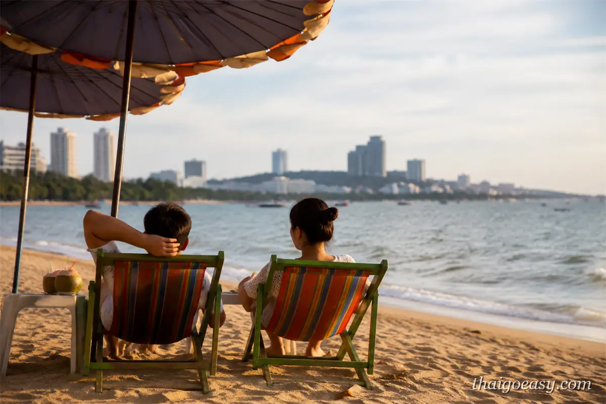 兩名旅客坐在芭達雅沙灘的條紋帆布椅上看海，旁邊小桌放著椰子，遠方是市區天際線