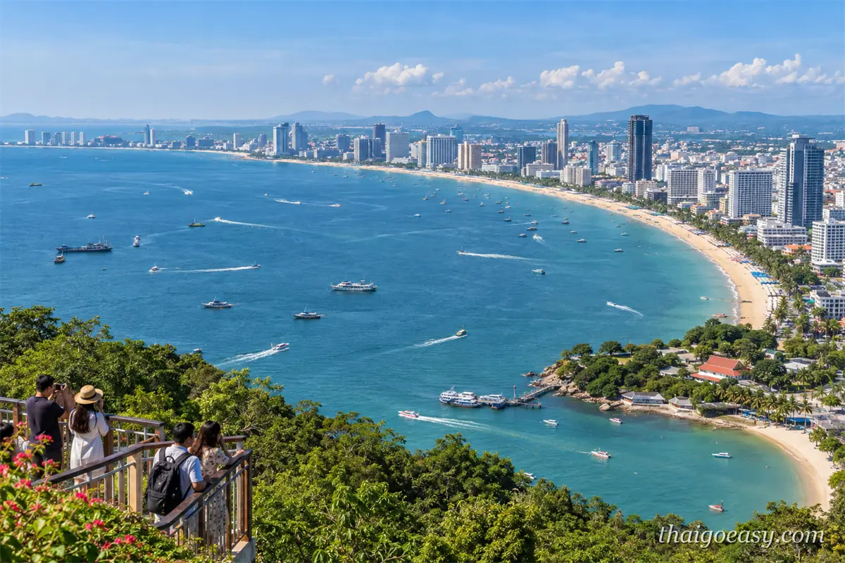 從山丘觀景台俯瞰芭達雅整條彎月形海灣，沿岸是成排高樓與弧形沙灘，海面散布快艇與客船，前景有遊客倚著欄杆拍照