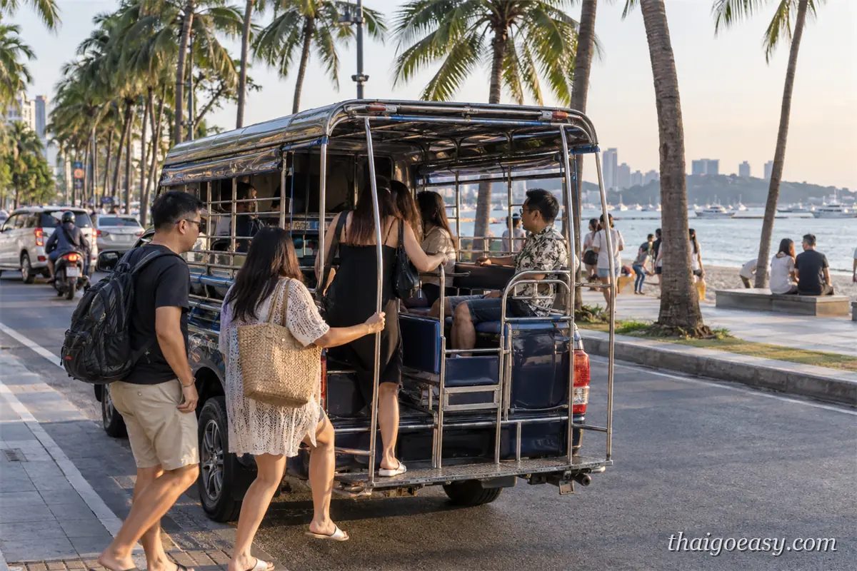 旅客正要登上停在芭達雅海濱路旁的雙條車，車廂是敞開式的雙排藍色座椅，路旁是椰子樹與沙灘