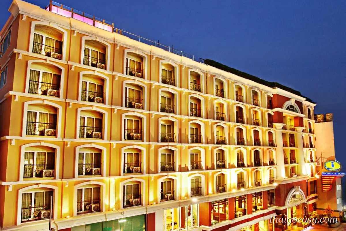 Intimate Hotel Pattaya 夜間外觀，米黃色歐式立面與逐層拱形陽台，右下角是飯店招牌
