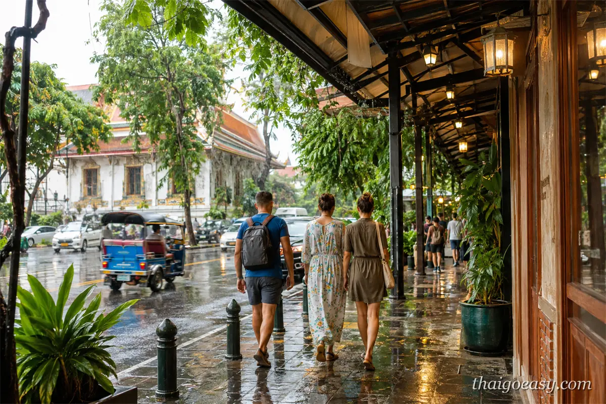 泰國雨季的曼谷街景，旅客走在騎樓下，路面積水映著店家燈光與嘟嘟車