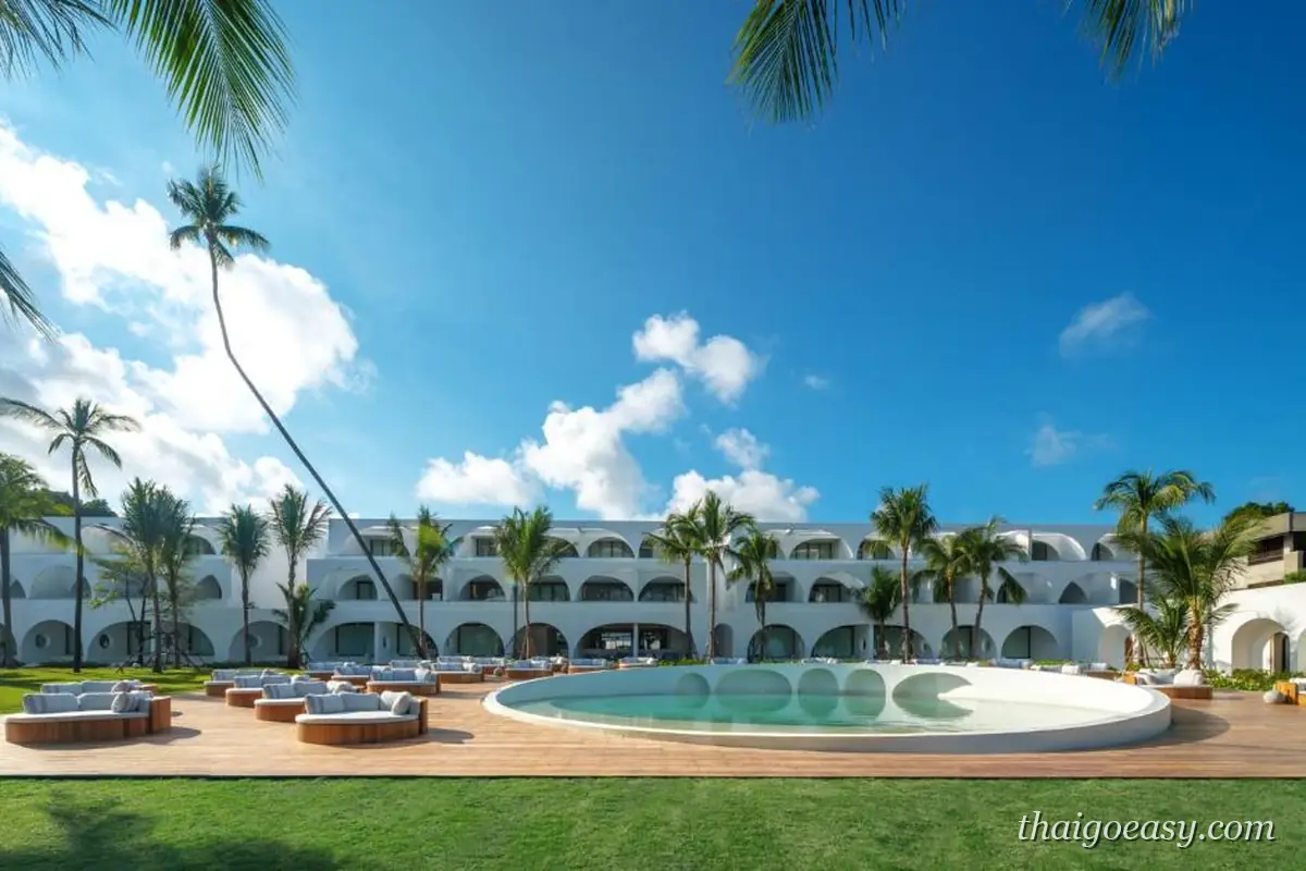 SALA Samui Chaweng Beach Resort 的全白拱廊建築圍著中庭，前方是曲線造型泳池、木平台躺椅與草坪，椰子樹斜伸入鏡