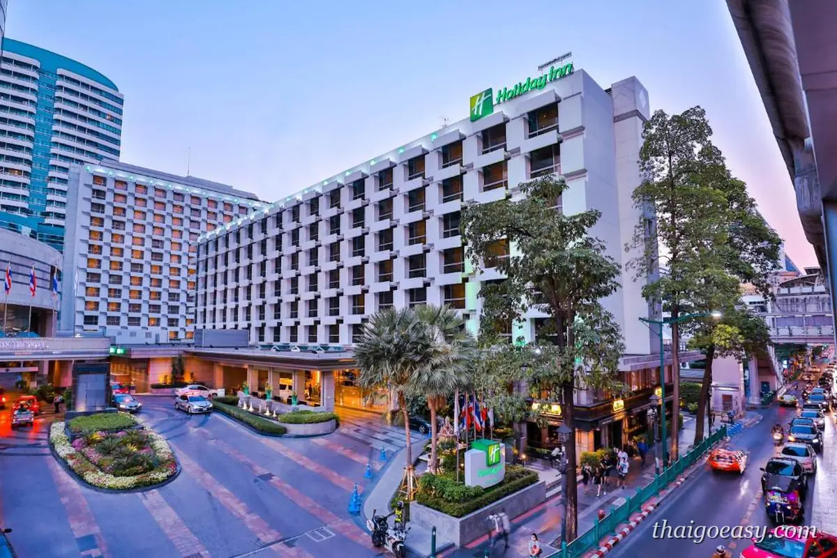 曼谷假日飯店黃昏外觀，屋頂掛著綠色 Holiday Inn 招牌，前方是門口車道與旗幟