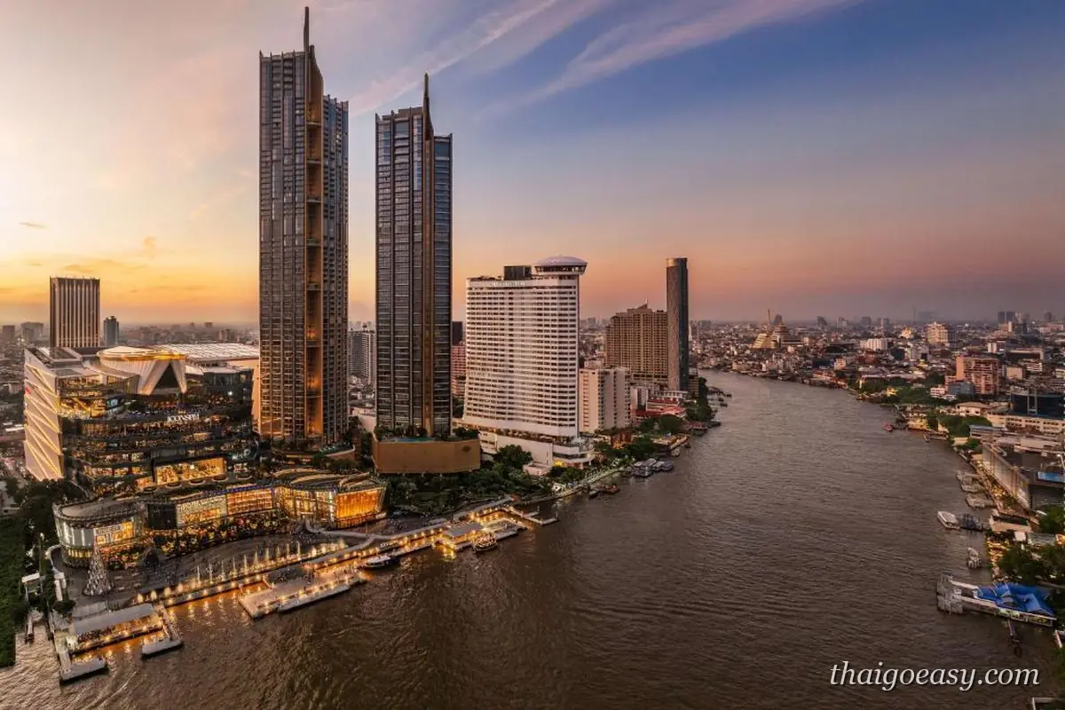 黃昏的昭披耶河畔俯瞰，中央是 Millennium Hilton Bangkok 的白色高樓與圓頂，左側是 ICONSIAM 商場與兩棟高塔，河岸碼頭亮著燈，右側是河對岸的城區