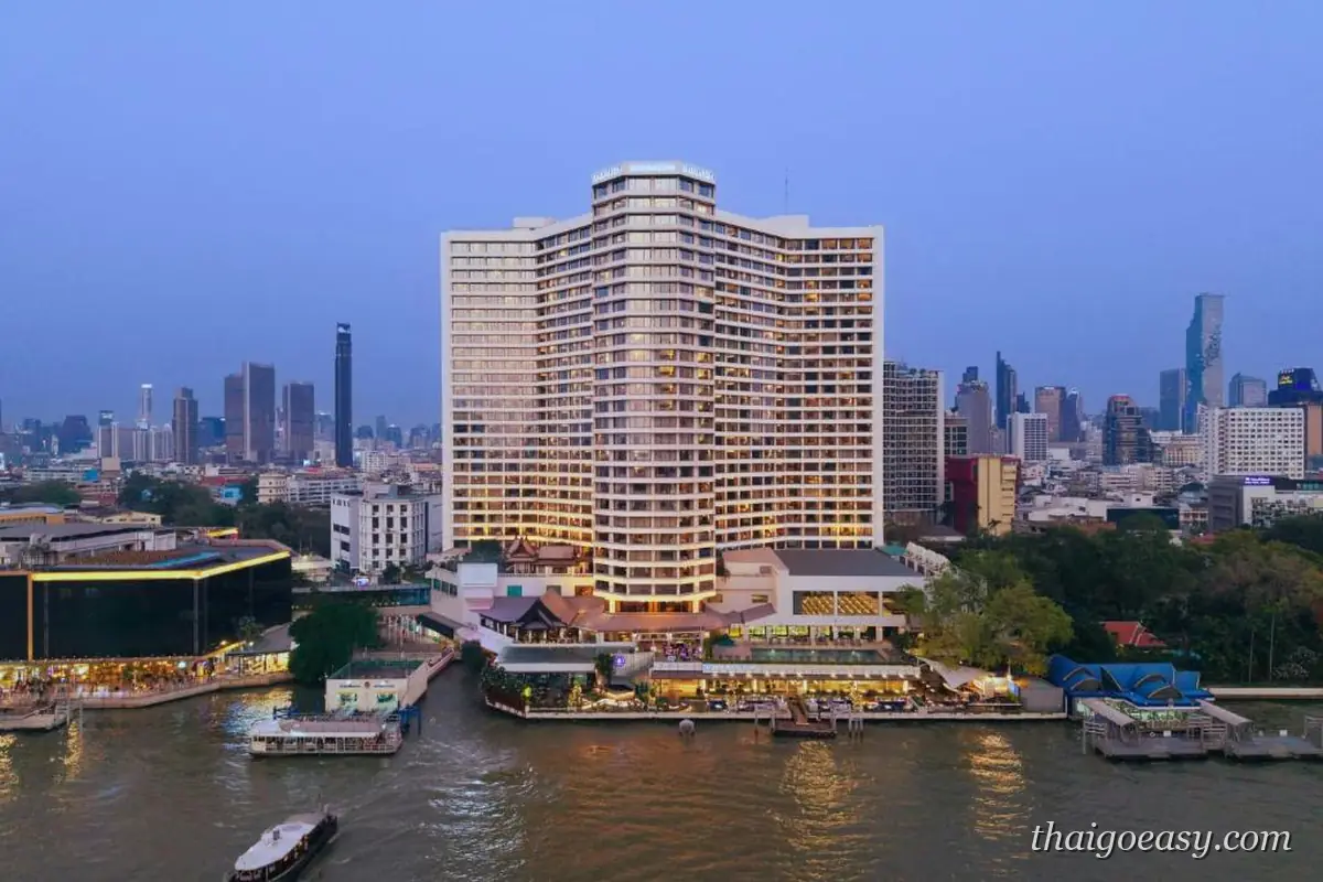 暮色中從河面望向 Royal Orchid Sheraton 的弧形高樓，樓頂亮著招牌、客房窗戶透出燈光，前方是河畔碼頭