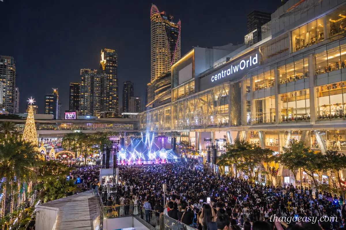 夜晚的 CentralWorld 前廣場，舞台打著藍紫色燈光、台前擠滿觀眾，左側立著點燈的聖誕樹，後方是亮燈的商場大樓