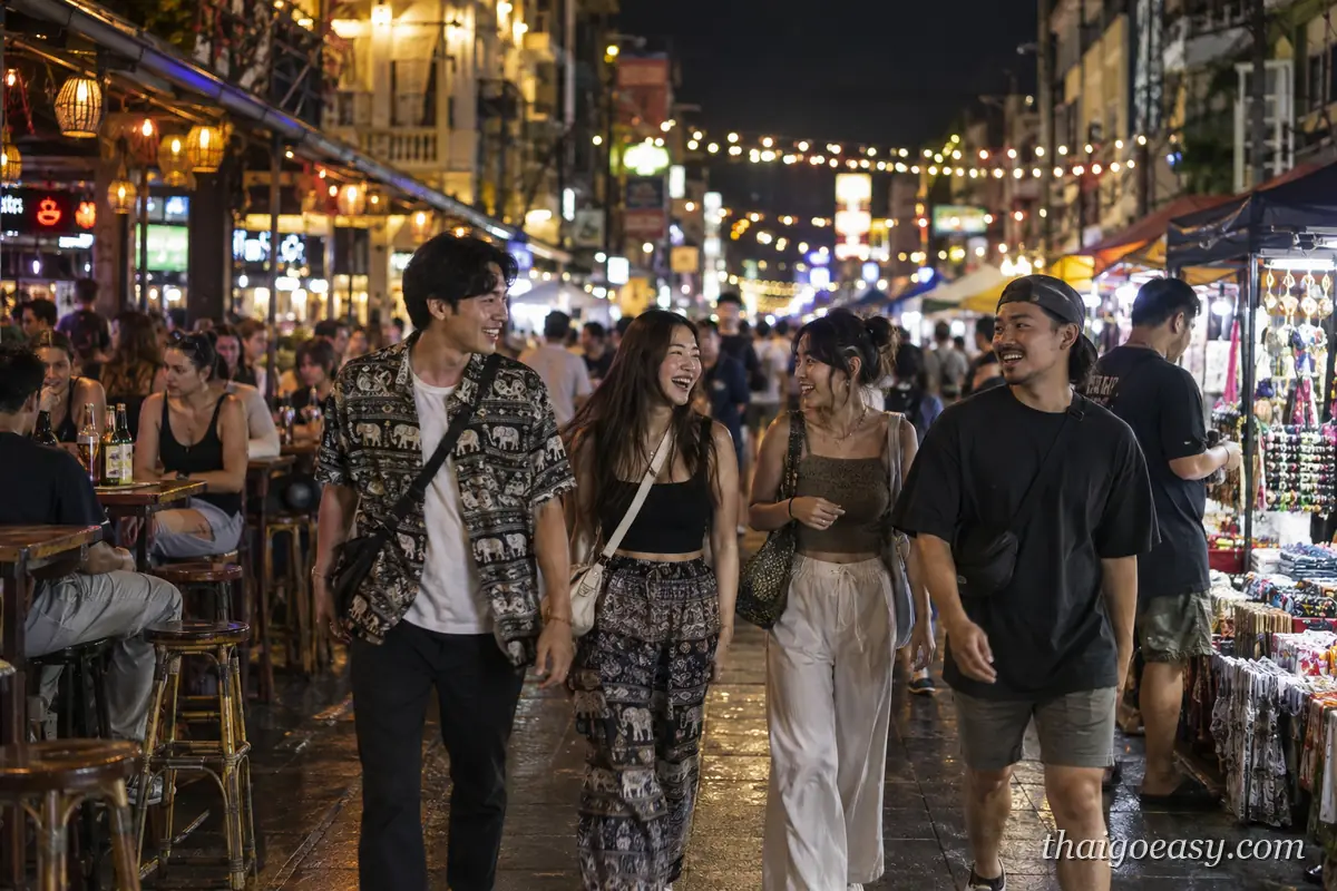 夜晚的考山路街頭，遊客與攤販擠在掛著串燈的街邊，前方一群年輕旅客笑著走過
